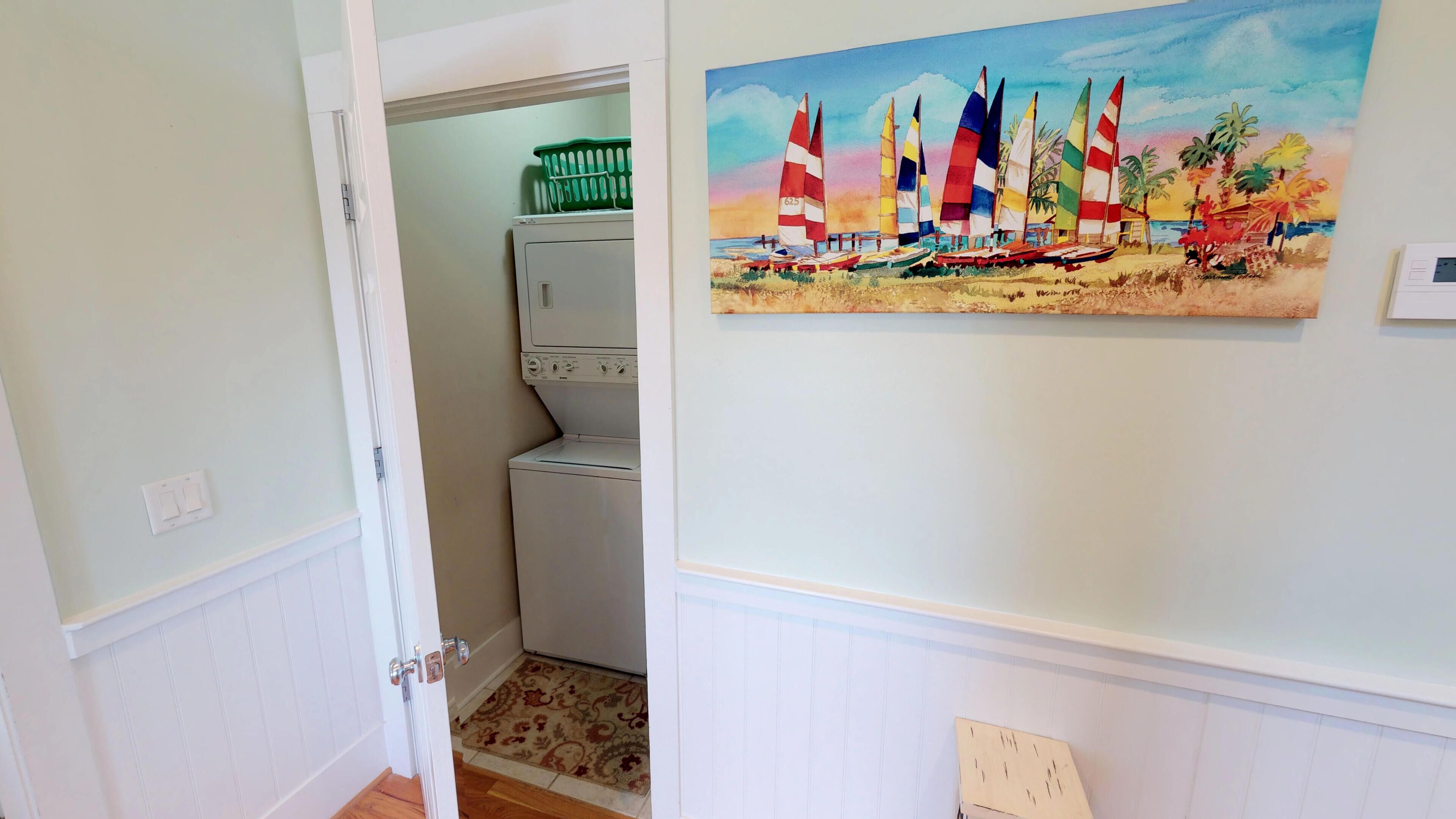 A hallway with a laundry area, a painting of sailboats on a beach, and a door leading to another room.