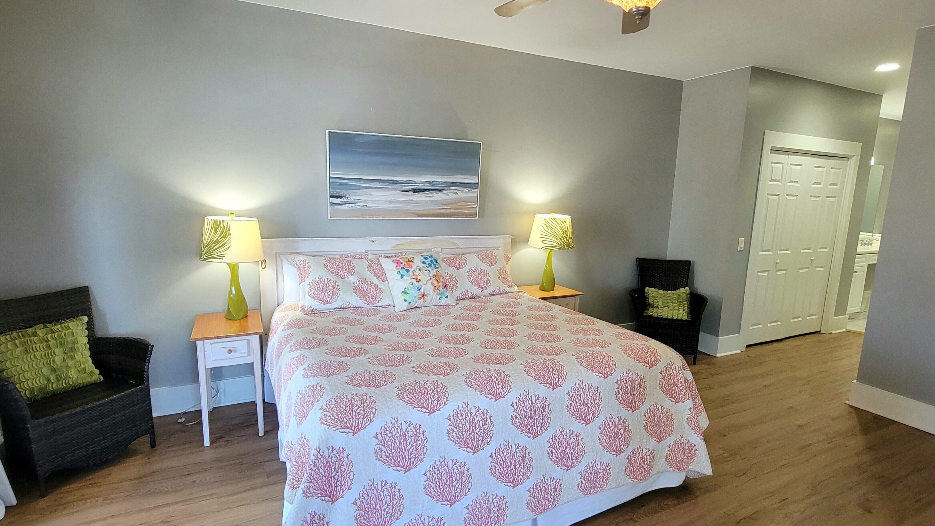 A bedroom with a bed, two bedside tables, a chair, a painting on the wall, and a door in the background.