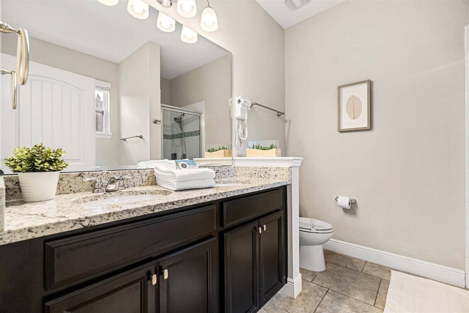 Une salle de bain dotée d’un comptoir en granit, de meubles sombres, de toilettes blanches et d’un grand miroir avec éclairage intégré.