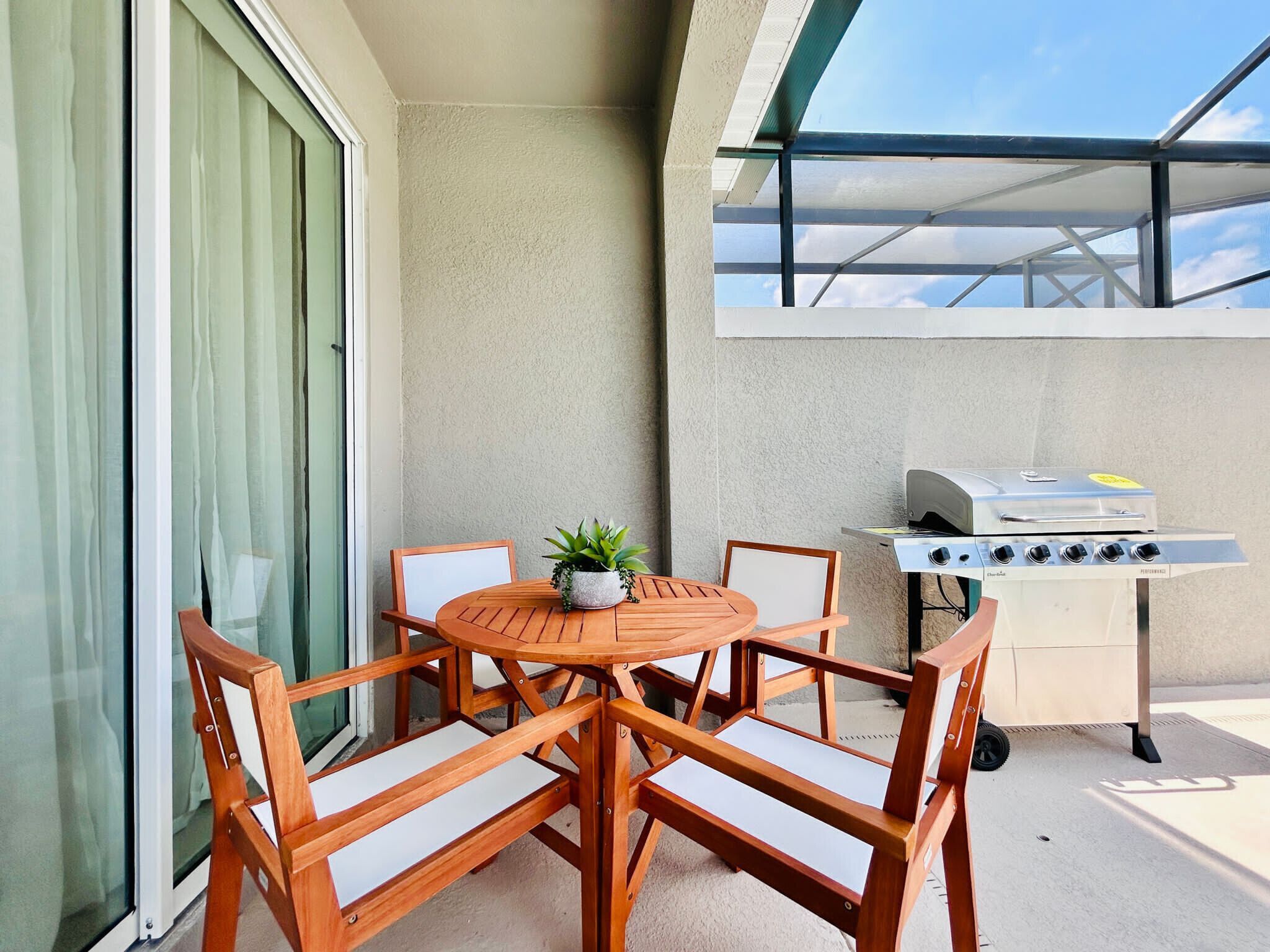 A balcony with a wooden table and chairs, a grill, and a view of the sky through a glass door.