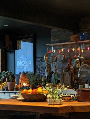A kitchen with a rustic design, featuring a wooden countertop, a variety of fruits and vegetables, and decorative items like a ceramic owl and hanging lights.