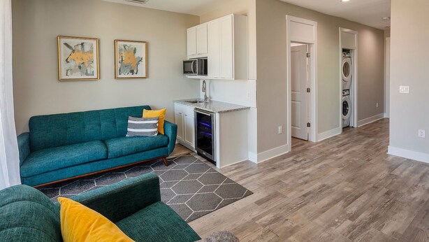 A modern living room with a teal sofa, a small kitchen area, and a hallway leading to a laundry room.