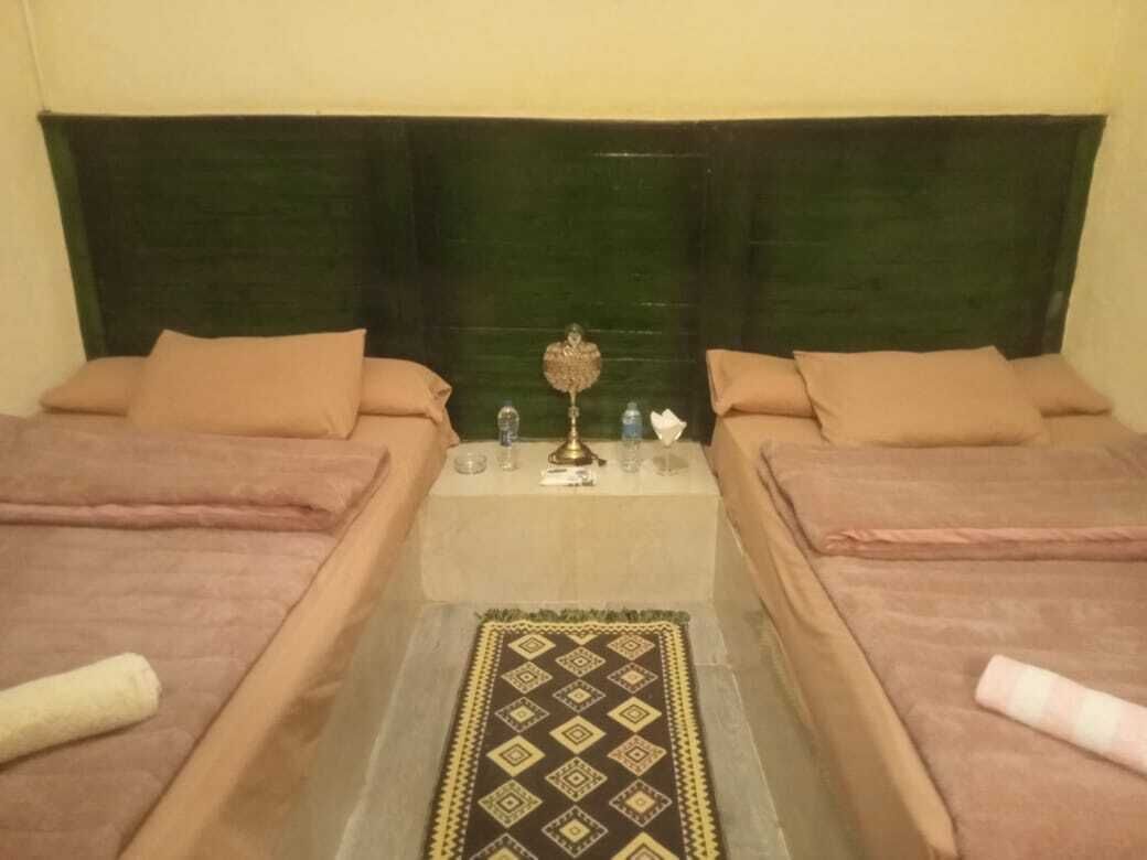 Una habitación con dos camas, una mesa central con lámpara y una alfombra estampada.
