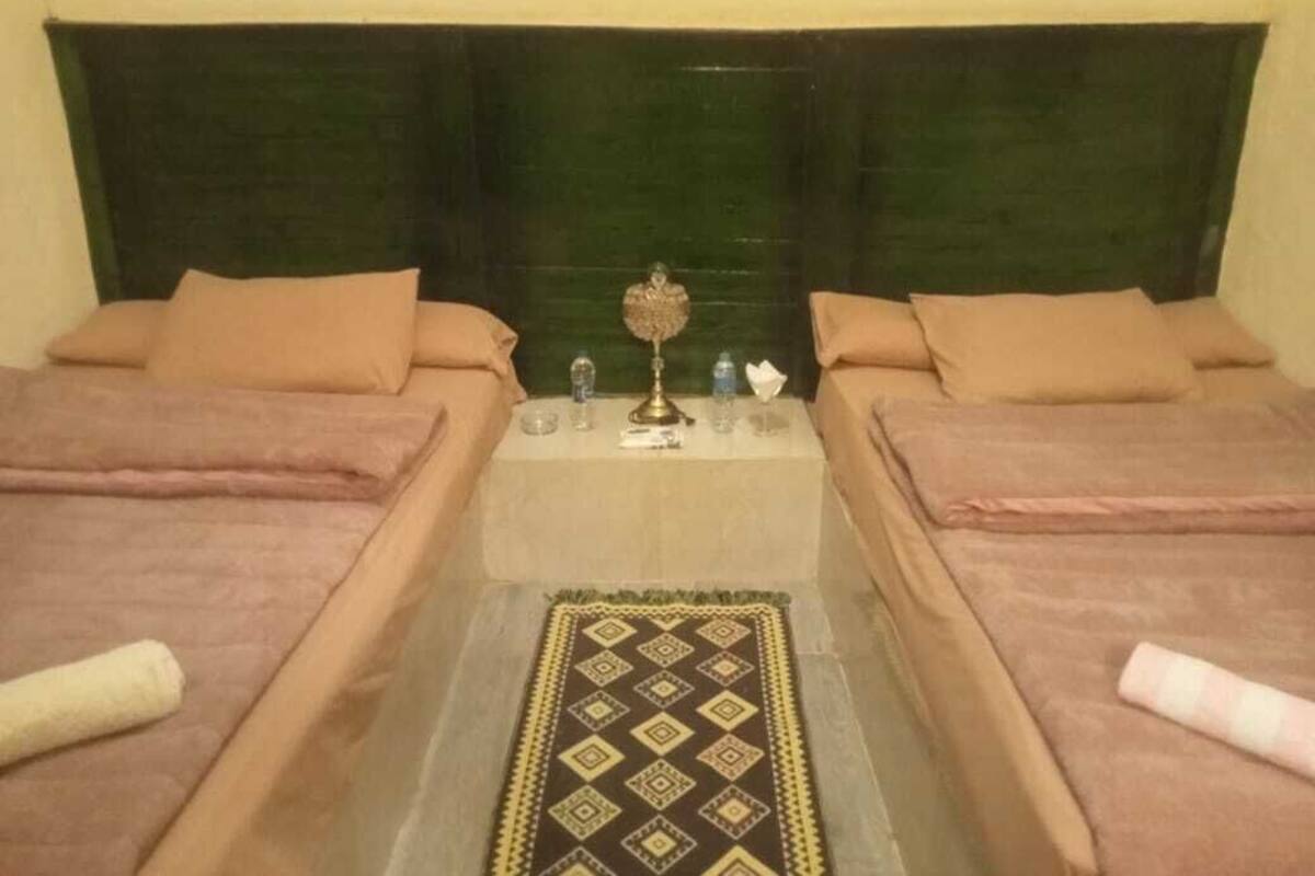 Una habitación con dos camas, una mesa central con lámpara y una alfombra estampada.
