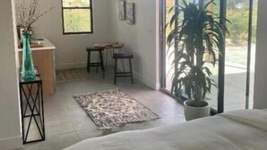 Une chambre avec un grand lit, une petite table, deux chaises, une plante en pot et une vue sur l’extérieur grâce à une grande fenêtre.