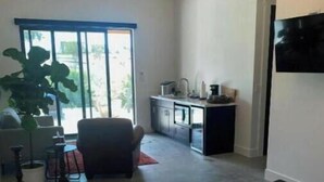 A modern hotel room with a large bed, a seating area with a chair and a table, a small kitchenette, and a large window with a view of greenery. - Villa Blanc, Private Individual Suite @ Hidden Hacienda (Temecula)