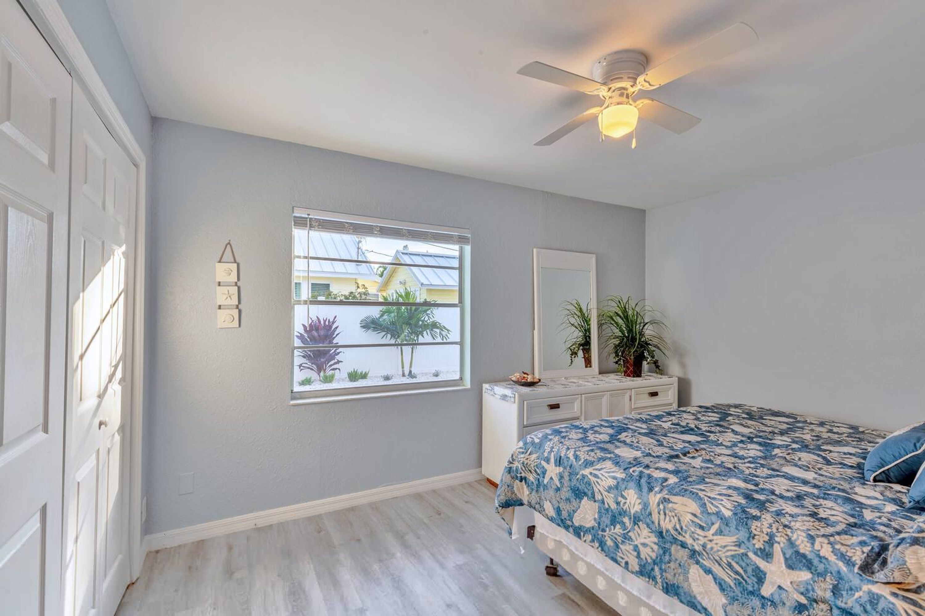 A bedroom with a bed, a ceiling fan, a window with a view of palm trees, and a dresser.