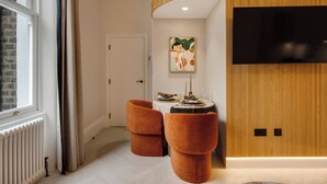 Un salon moderne avec un téléviseur fixé sur un mur recouvert de panneaux en bois, un coin repas avec une petite table et des chaises, et une grande fenêtre avec des rideaux.