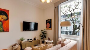 A living room with a beige sofa, a television, a plant, and a window with a view of buildings and trees. - Notting Hill | 1-Bedroom Deluxe Apartment (London)