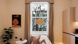A cozy living room with a sofa, a small side table with a book, a potted plant, and a window with curtains. - Notting Hill | 1-Bedroom Deluxe Apartment (London)