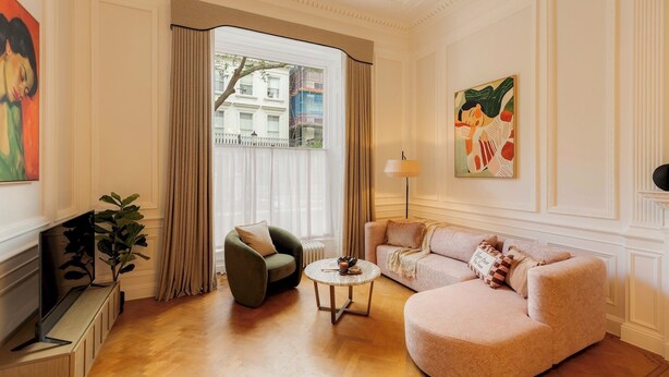 Un salon avec un canapé, un fauteuil et une petite table ronde. Il y a une fenêtre avec des rideaux, un radiateur et un tableau au mur.