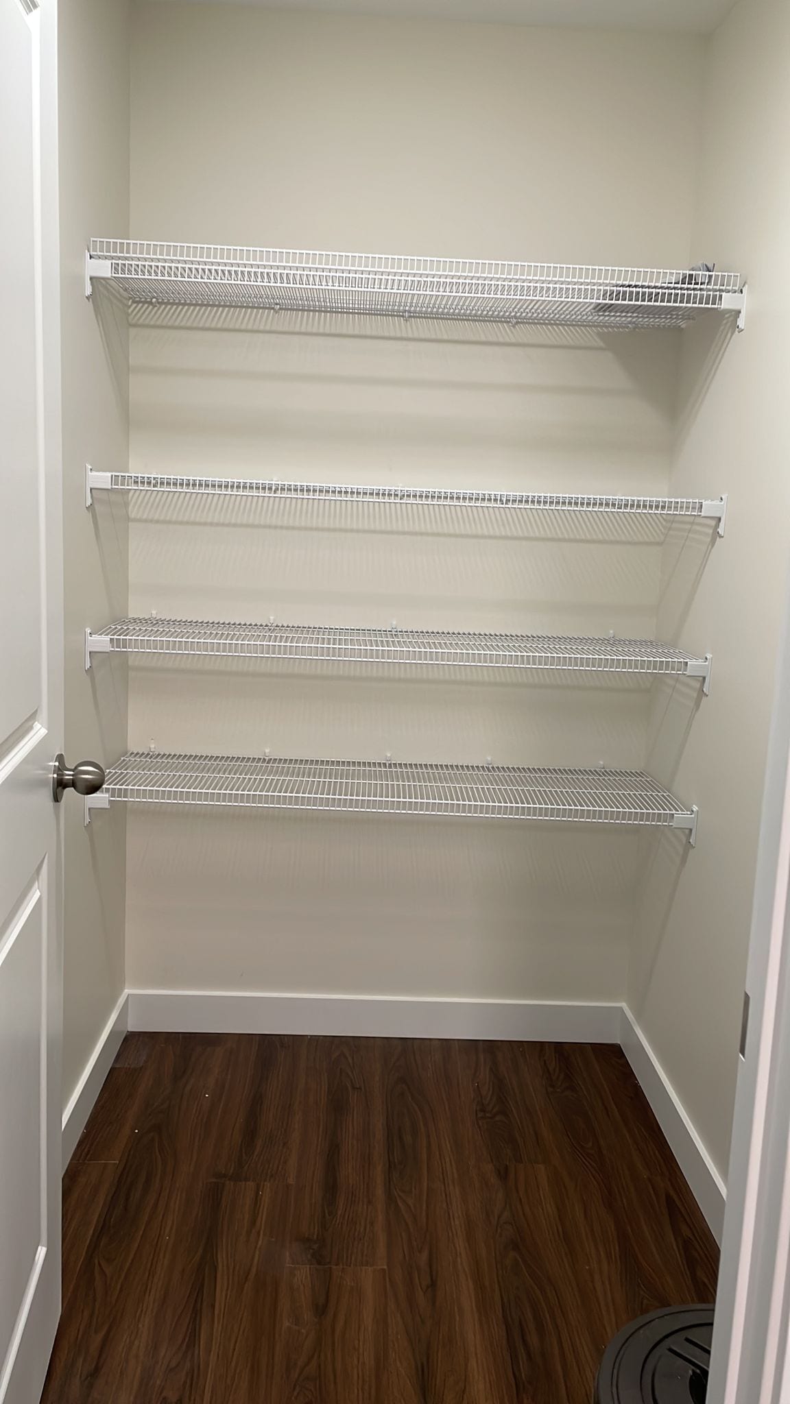 A closet with four white wire shelves against a white wall, wooden flooring, and a door with a round handle.