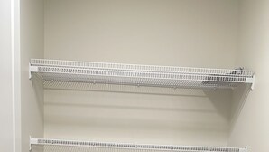 Un armario con cuatro estantes metálicos blancos sobre un suelo de madera, y una puerta con pomo redondo.
