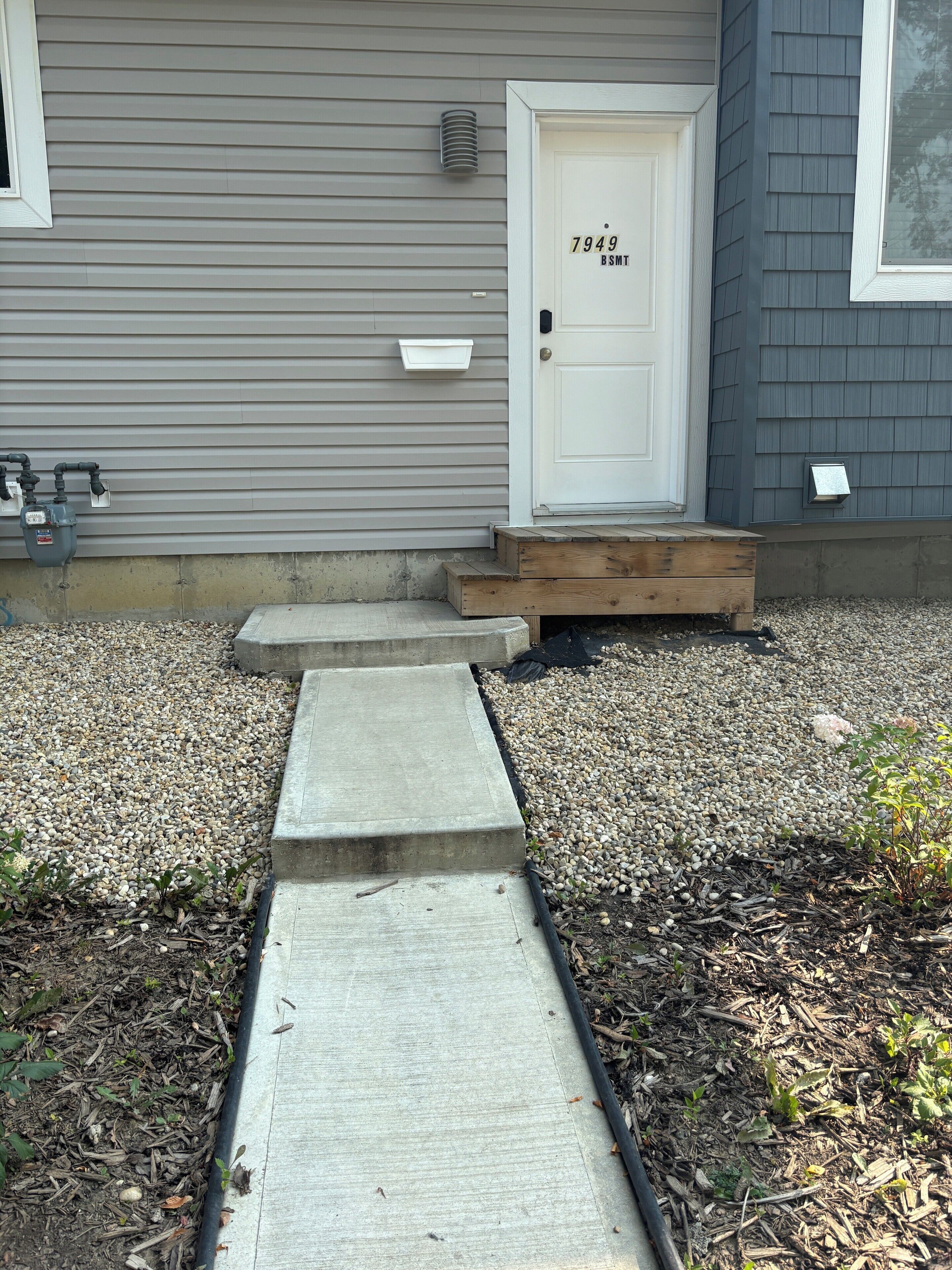 A house with a grey siding exterior, a white front door with the number 7949, and a wooden front step leading to the entrance.