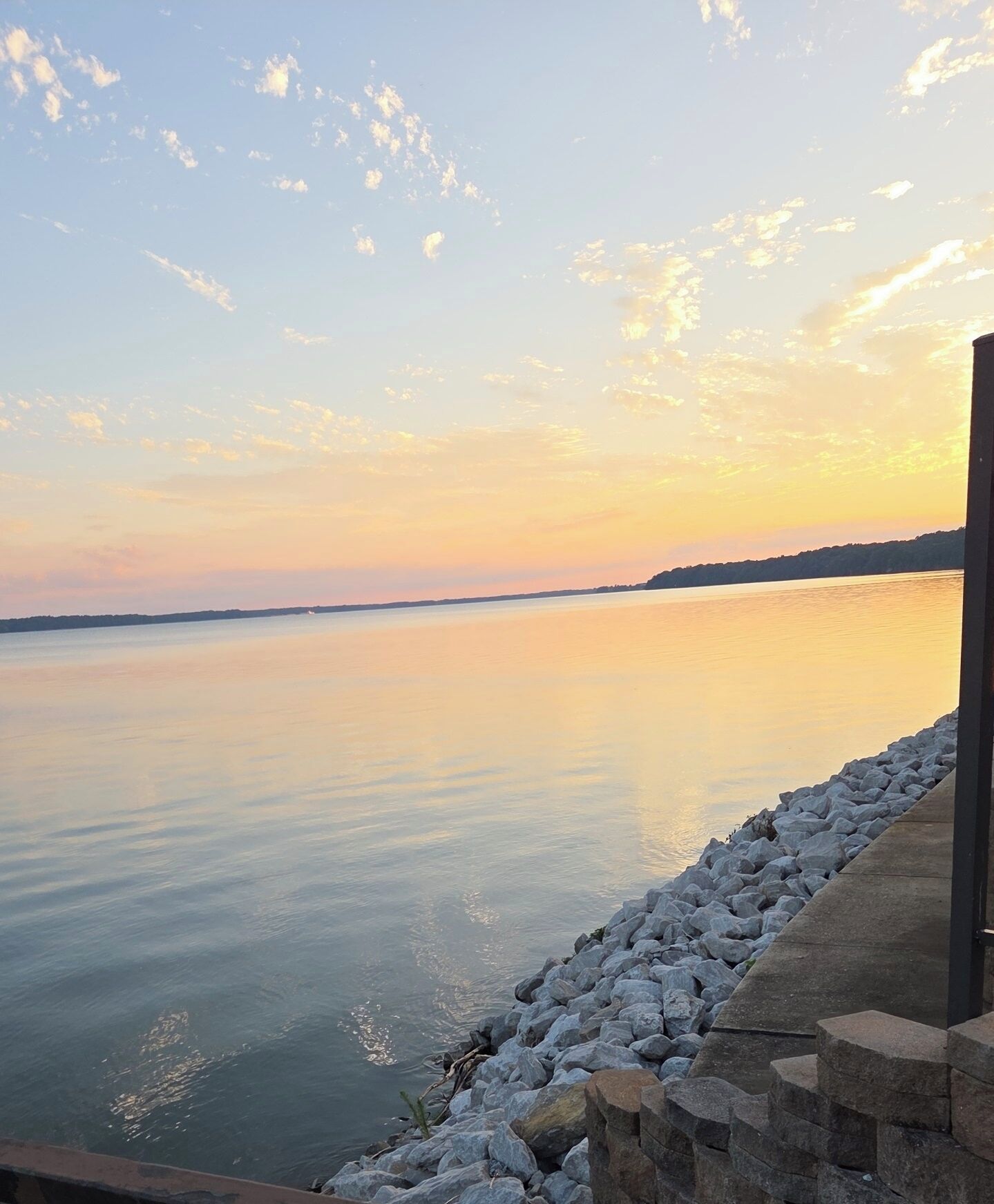 A calm body of water with a rocky shoreline and a distant landmass under a clear sky.