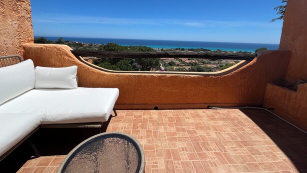 A balcony with a white sofa and a glass table, overlooking a coastal view with houses and the sea. - Villa Aurea Snow White 139 (Costa Rei)