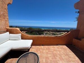 Un balcone con un divano bianco e un tavolo in vetro, vista sulla costa, case e mare.
