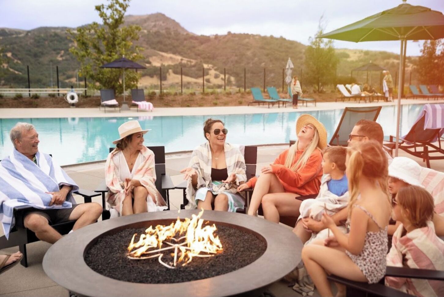 Eine Gruppe von Personen sitzt um eine runde Feuerstelle am Pool, im Hintergrund liegen Liegestühle und Sonnenschirme.