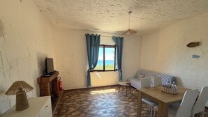 A room with a wooden floor, a sofa, a dining table with chairs, a TV on a stand, and a window with curtains.