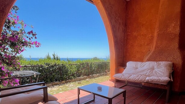 A room with a bed, a small table, and a chair, with a view of the sea and greenery outside.