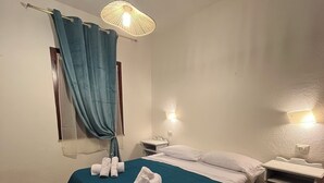 A small, tidy room with a bed, a nightstand, a wall-mounted lamp, and a window with curtains.