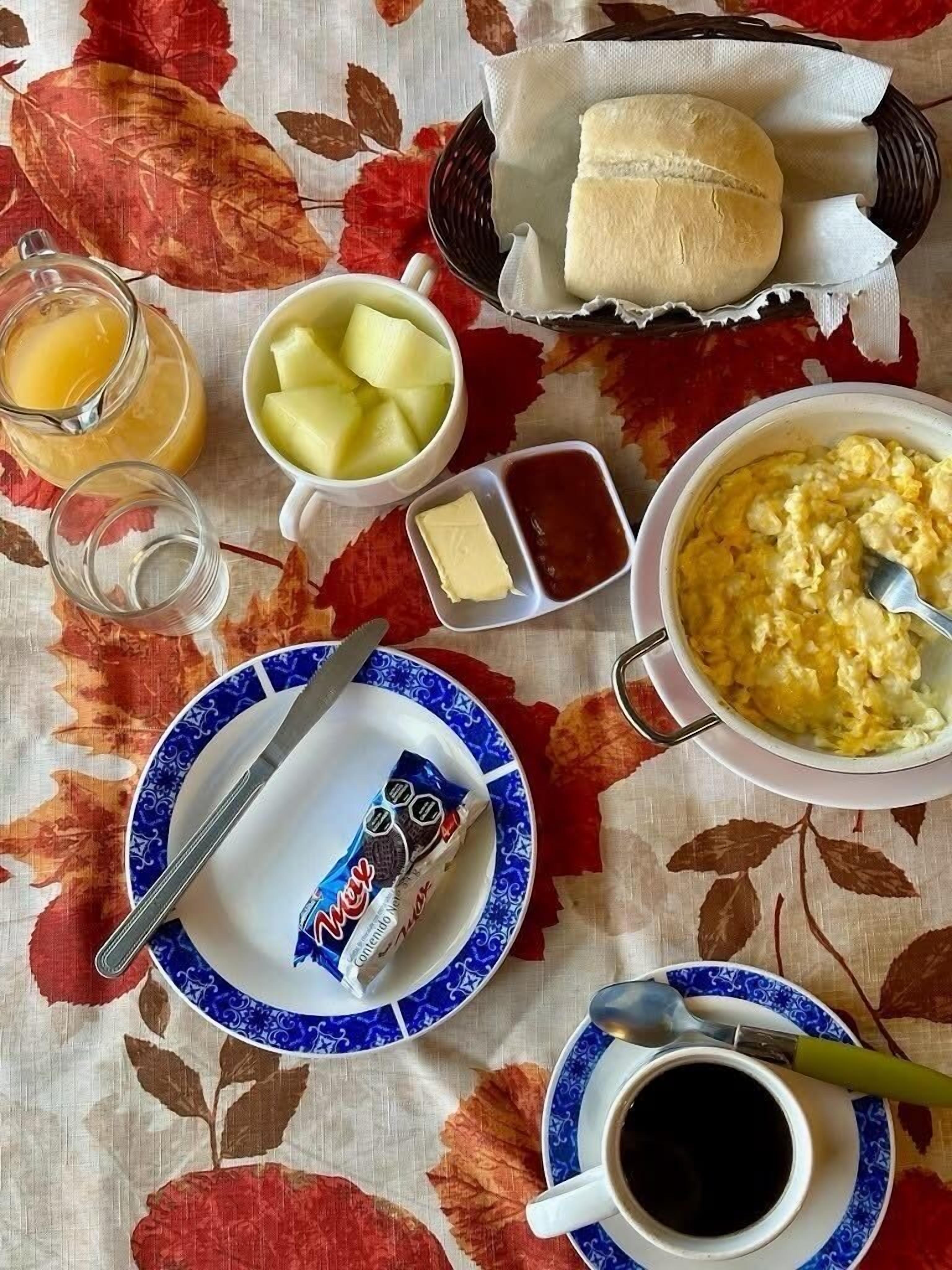 Et morgenbord med et fad med røræg, en skål med frugt, et glas appelsinjuice, en kurv med to boller, en lille skål smør og en kop kaffe.