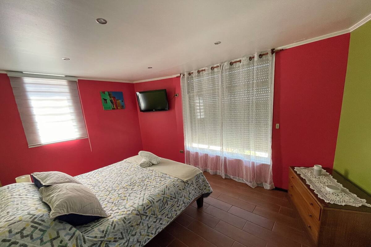 Une chambre à coucher avec un lit, un téléviseur fixé au mur, une fenêtre avec des stores et une commode en bois.