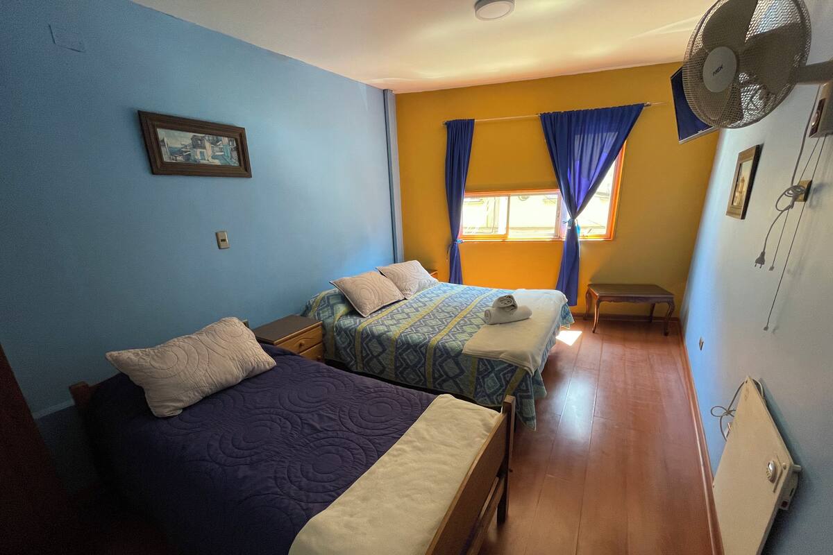 Une chambre avec deux lits, une petite table, un ventilateur sur pied et une fenêtre avec des rideaux.