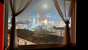 Vista de un puerto con grúas y contenedores marítimos a través de una ventana con cortinas traslúcidas.