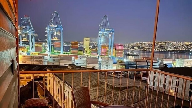 Un balcón con mesa y sillas, con vista a un puerto con contenedores marítimos y grúas.