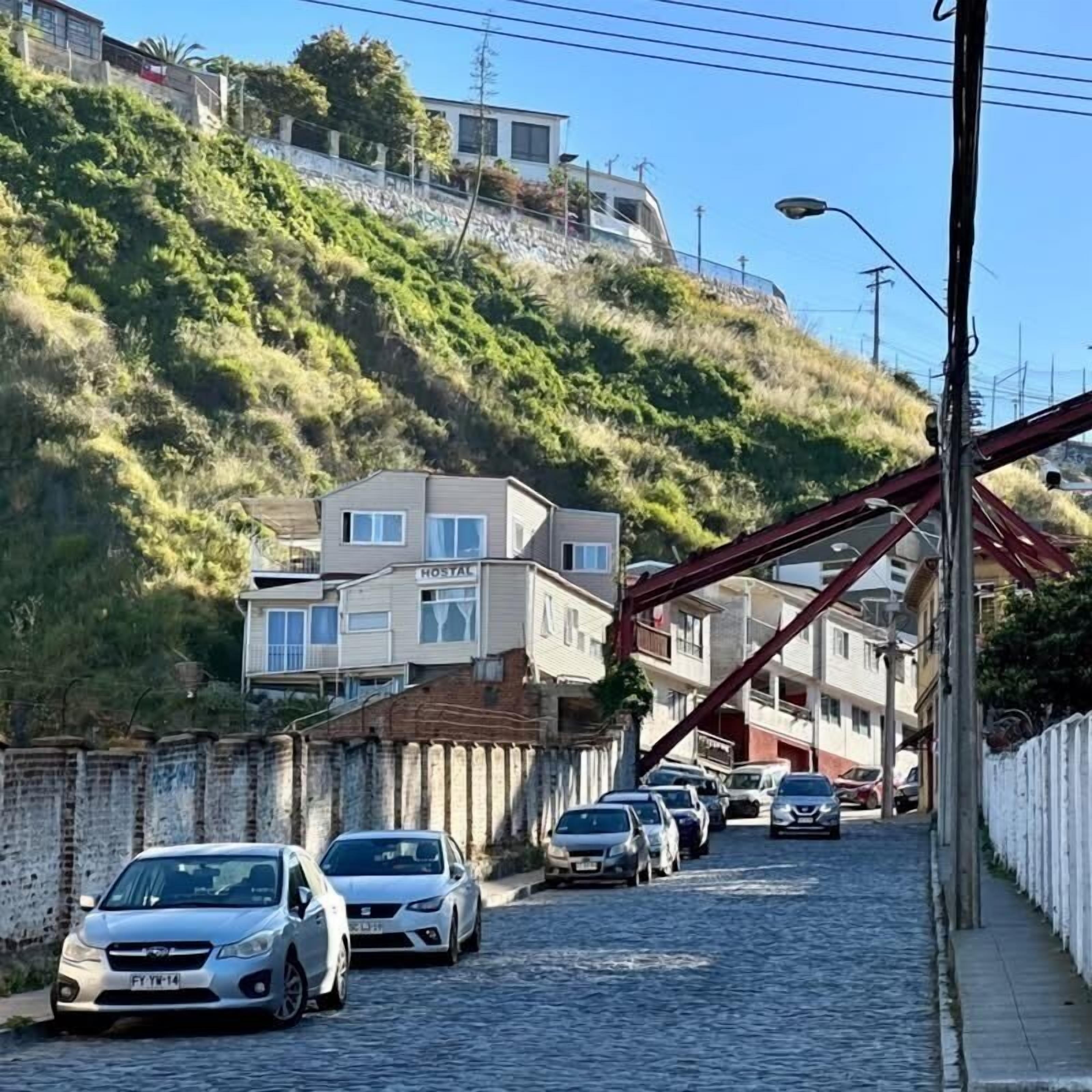 Un hotel llamado "HOSTAL" en una ladera, con una carretera pavimentada y coches aparcados.