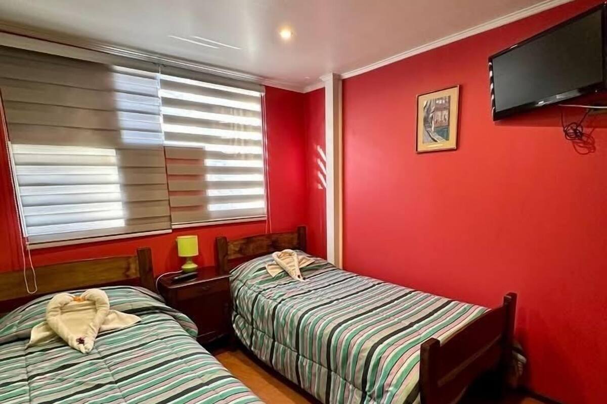 Une chambre avec deux lits, un téléviseur fixé au mur et une fenêtre avec des stores.