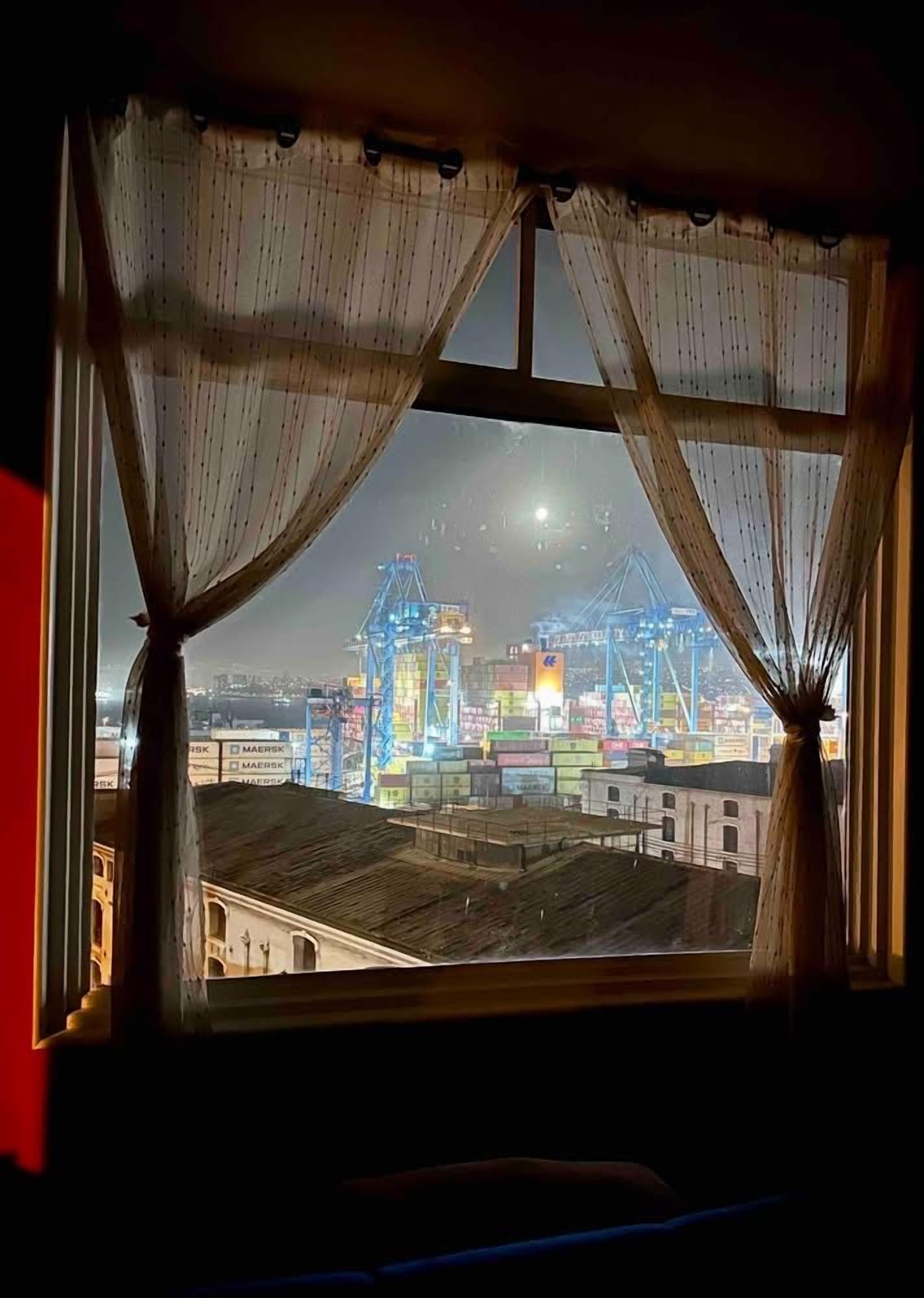 A view of a port with cranes and shipping containers through a window with sheer curtains.