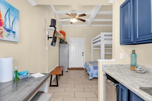 Une cuisine de taille réduite, avec un banc en bois, un ventilateur de plafond et une porte donnant sur une chambre.