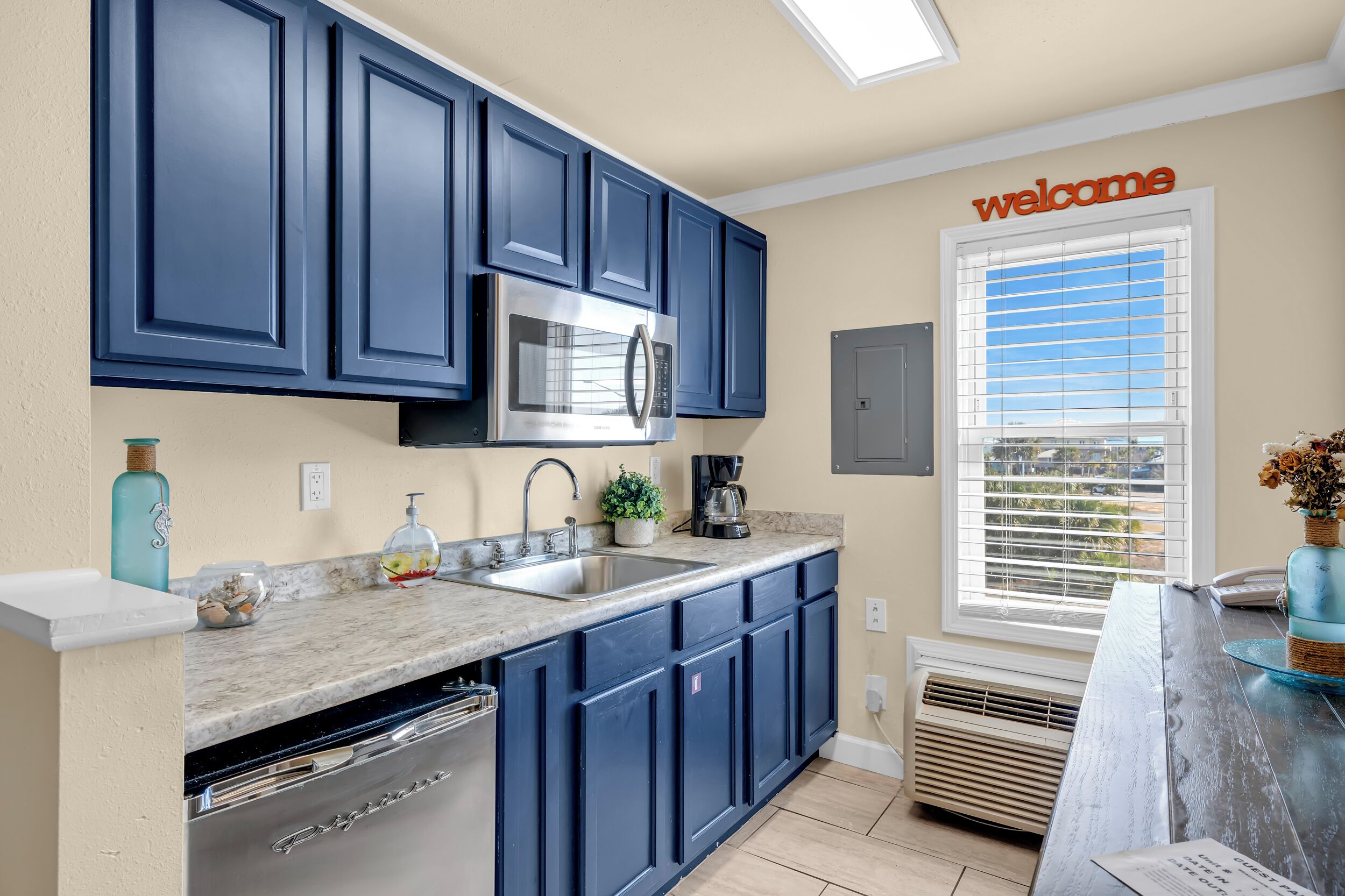 A kitchen with dark blue cabinets, a stainless steel microwave, a sink with a faucet, a coffee maker, and a window with blinds.
