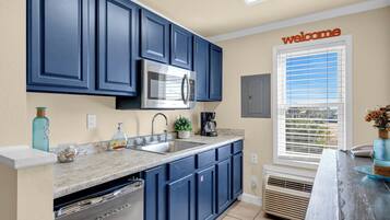 A kitchen with dark blue cabinets, a stainless steel microwave, a sink with a faucet, a coffee maker, and a window with blinds.