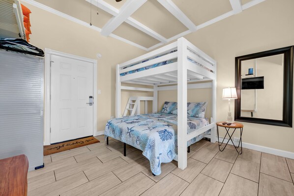Una cama litera con colcha azul estampada, una puerta blanca, una mesita con lámpara y un espejo en la pared.