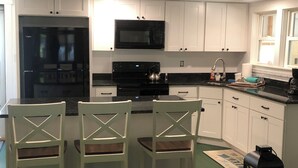 A kitchen with white cabinets, a black refrigerator, a microwave, and a stove. There are three chairs with wooden backs and seats.