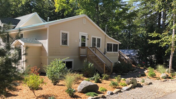 A two-story house with a wooden staircase leading to a deck, surrounded by a landscaped garden with various plants and rocks.