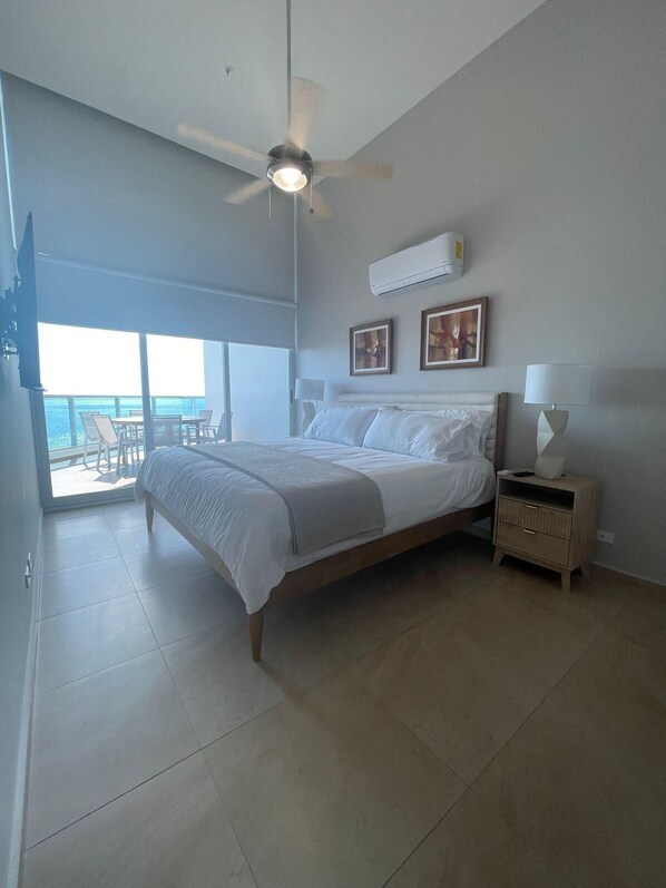 Un dormitorio con cama, mesita de noche, ventilador de techo y una ventana grande con vista al océano.