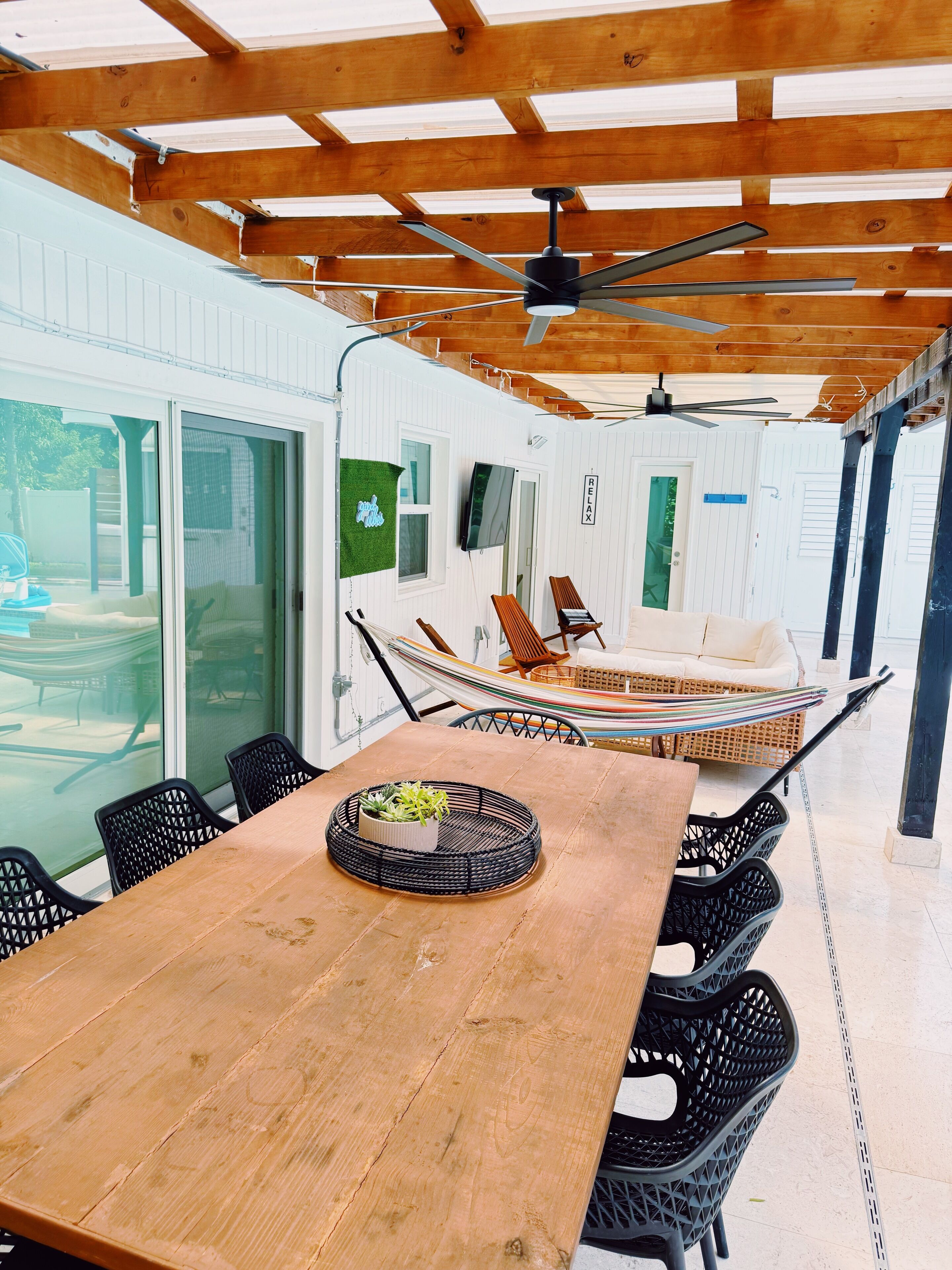 An outdoor patio with a wooden table, black chairs, a hammock, and a ceiling fan.