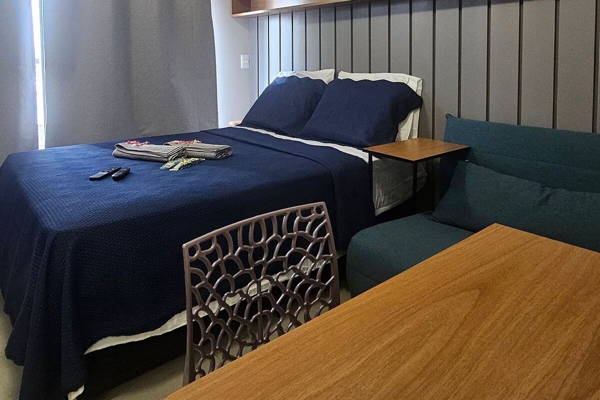A hotel room with a bed, a wooden table, a sofa, and a headboard with built-in storage.