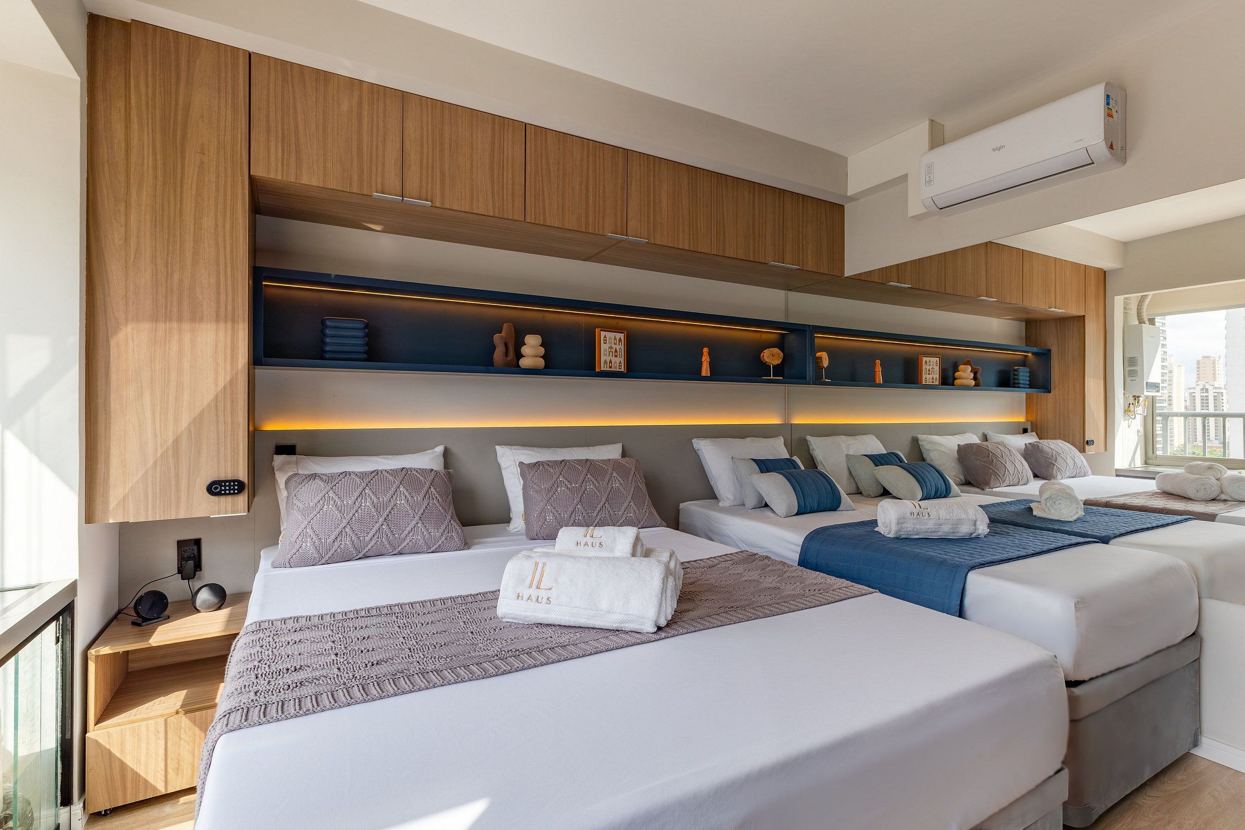 Une chambre d’hôtel moderne avec deux lits, une étagère intégrée agrémentée d’objets décoratifs, une table de chevet avec un téléphone et un haut-parleur, et un appareil de climatisation.