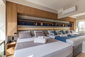 A modern hotel room with two beds, a built-in shelf with decorative items, a bedside table with a phone and speaker, and an air conditioning unit. - Condominio Helbor B.liv (São Paulo)