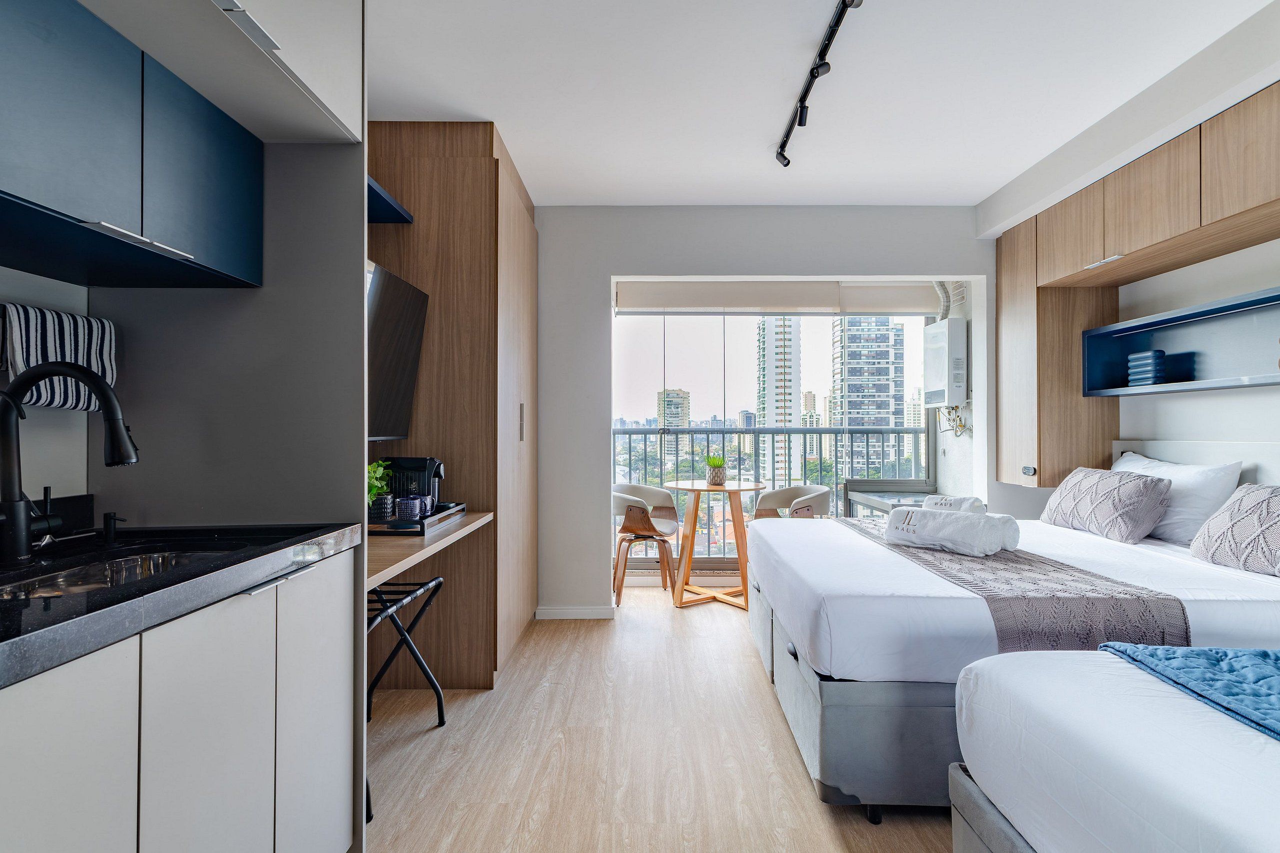 A modern hotel room with a kitchen area, a balcony with a city view, and a bed with a blue blanket.