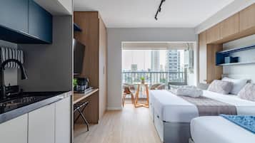 Quarto de hotel moderno com cozinha, varanda e vista para a cidade, e cama com manta azul.
