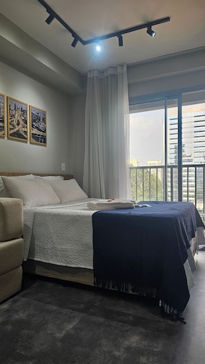 A bedroom with a bed, a chair, a balcony with a view, and three framed pictures on the wall. - Condominio Helbor B.liv (São Paulo)