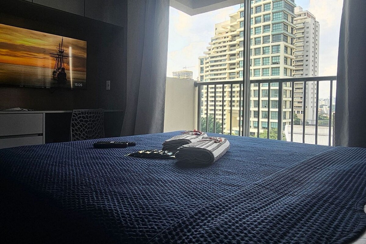 A hotel room with a bed, a TV, a window with a view of high-rise buildings, and a balcony.