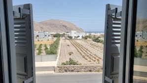 A view through an open window of a coastal town with a prominent white building, a rocky hillside, and a clear sky. - Leon Lux Apartment, quadruple room with private bathroom. (Emporio)