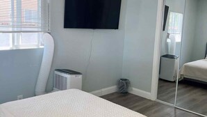 A bedroom with a bed, a ceiling fan, a TV mounted on the wall, a window with blinds, and a mirror in the adjacent room. - Studio Near Disney (Santa Ana)